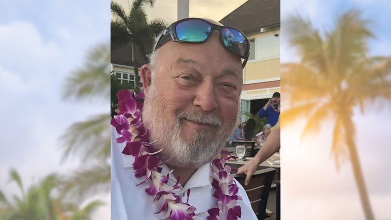 Donald F. Mlostek smiling with a Hawaiian lei around his neck, sunglasses on top of his head, and palm trees in the background.