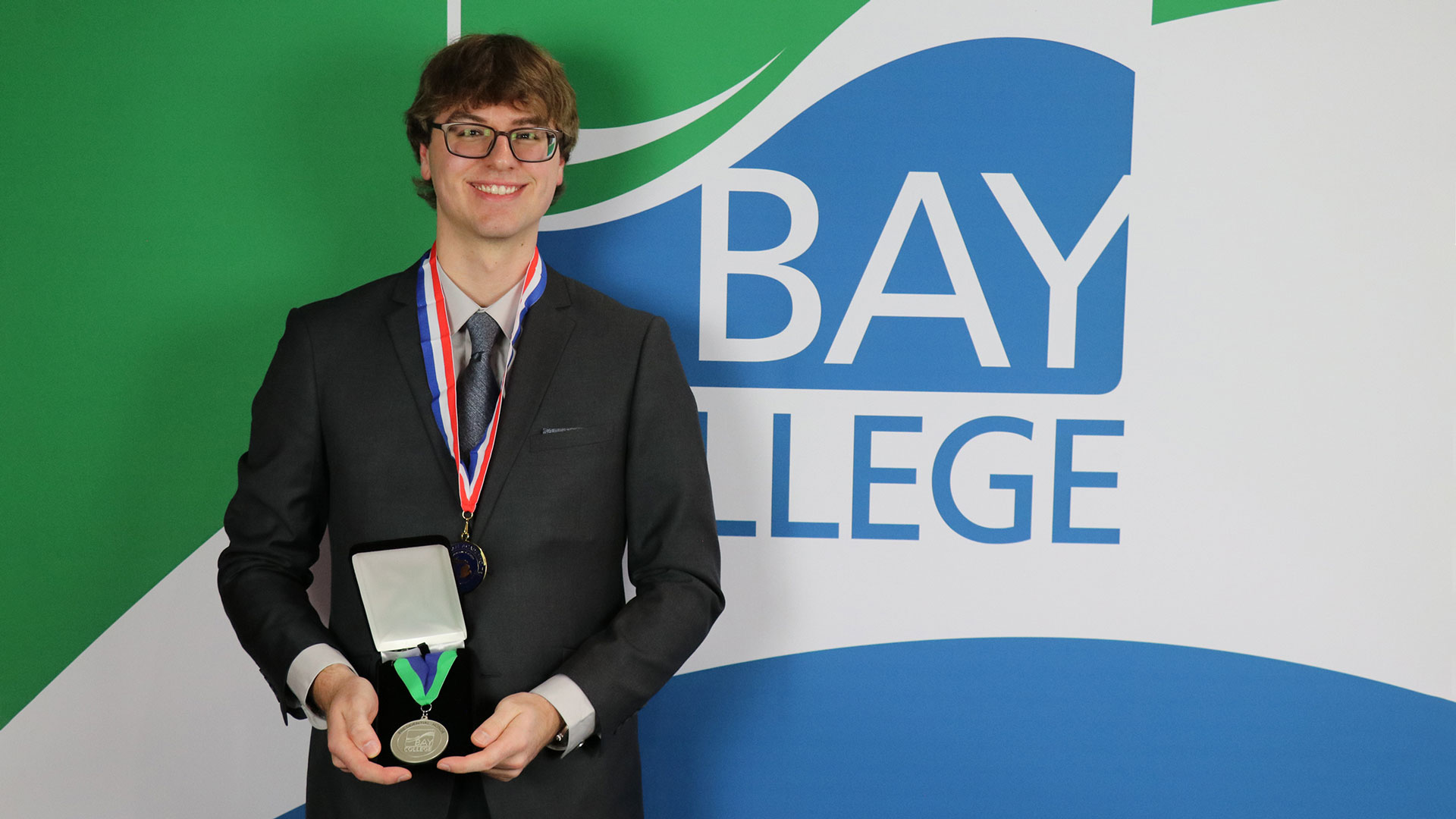 Gabriel Viau smiles in a charcoal suit holding his Presidential Honor medallion while wearing his All-Michigan medallion around his neck.