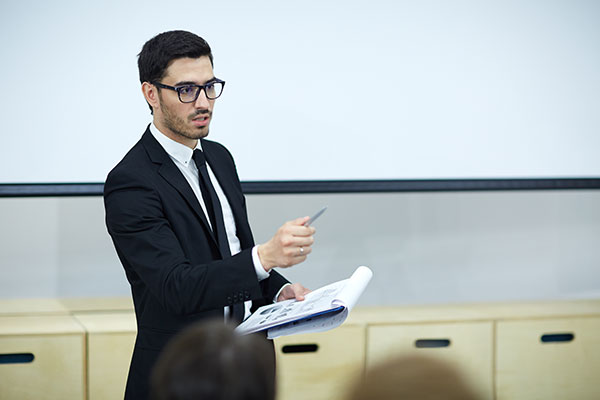 Man in a black suit and tie addressing a room of people